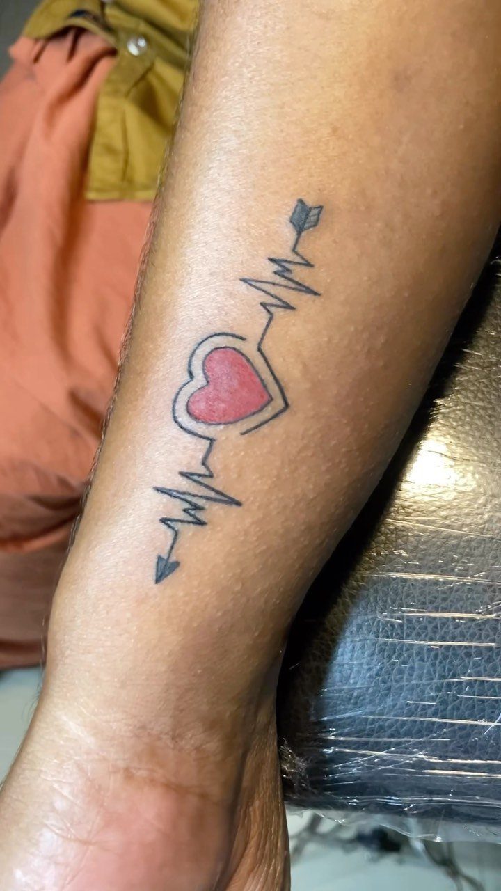 Heartbeat line tattoo with a red heart and arrow on a forearm.