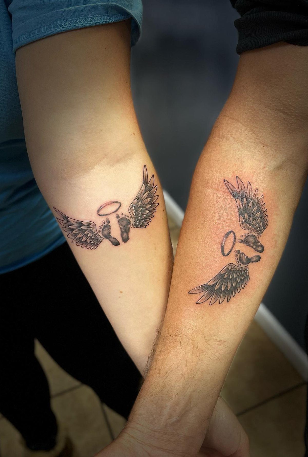 Matching tattoos of baby footprints with angel wings and halos on two forearms.