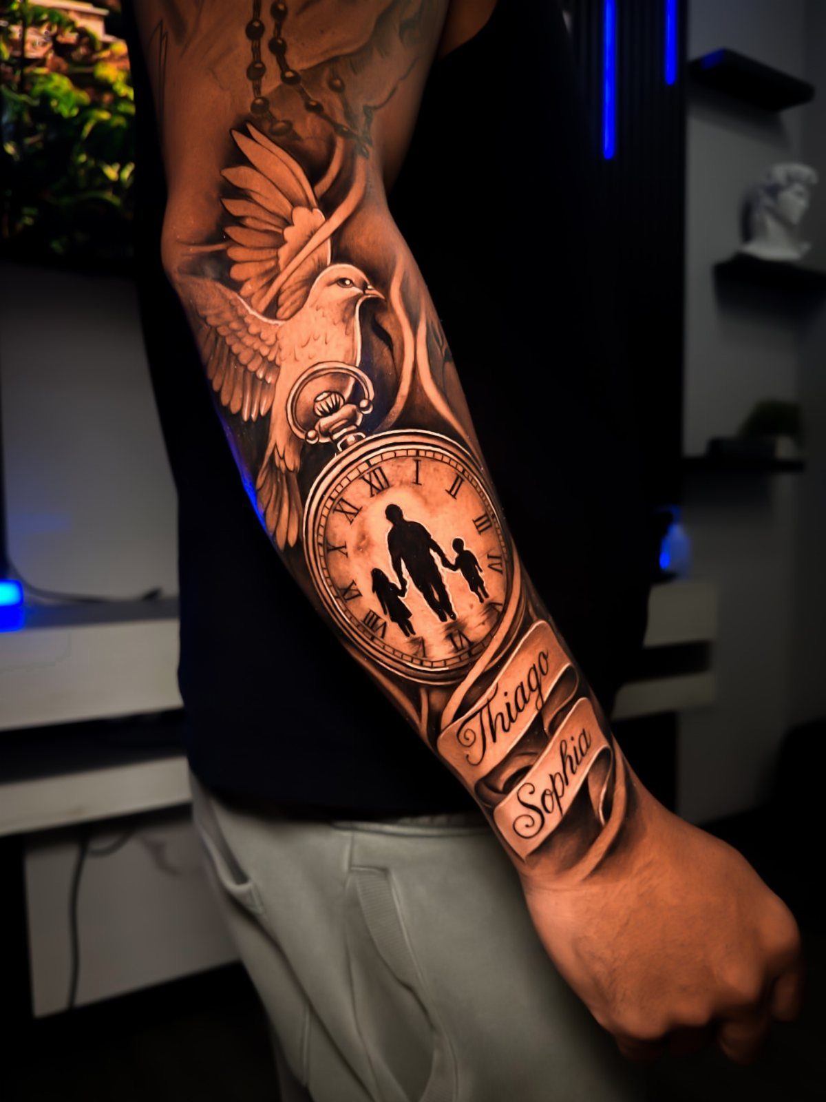 Pocket watch tattoo with family silhouette, a dove, and names on a forearm.
