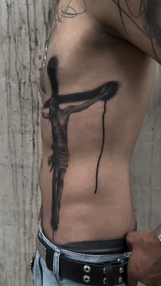 Crucifix tattoo on a person's side stomach and ribs in black and grey ink