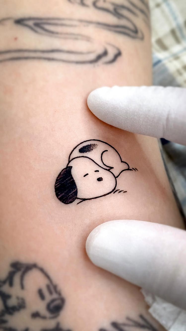 Cartoon tattoo of a small sleeping dog on skin, framed by gloved fingers.