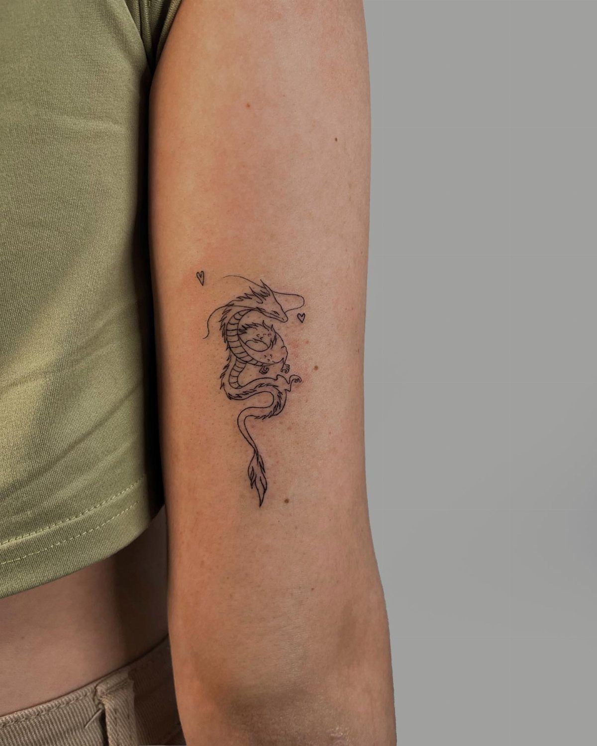 Small black ink dragon tattoo on a person's upper arm with tiny hearts.