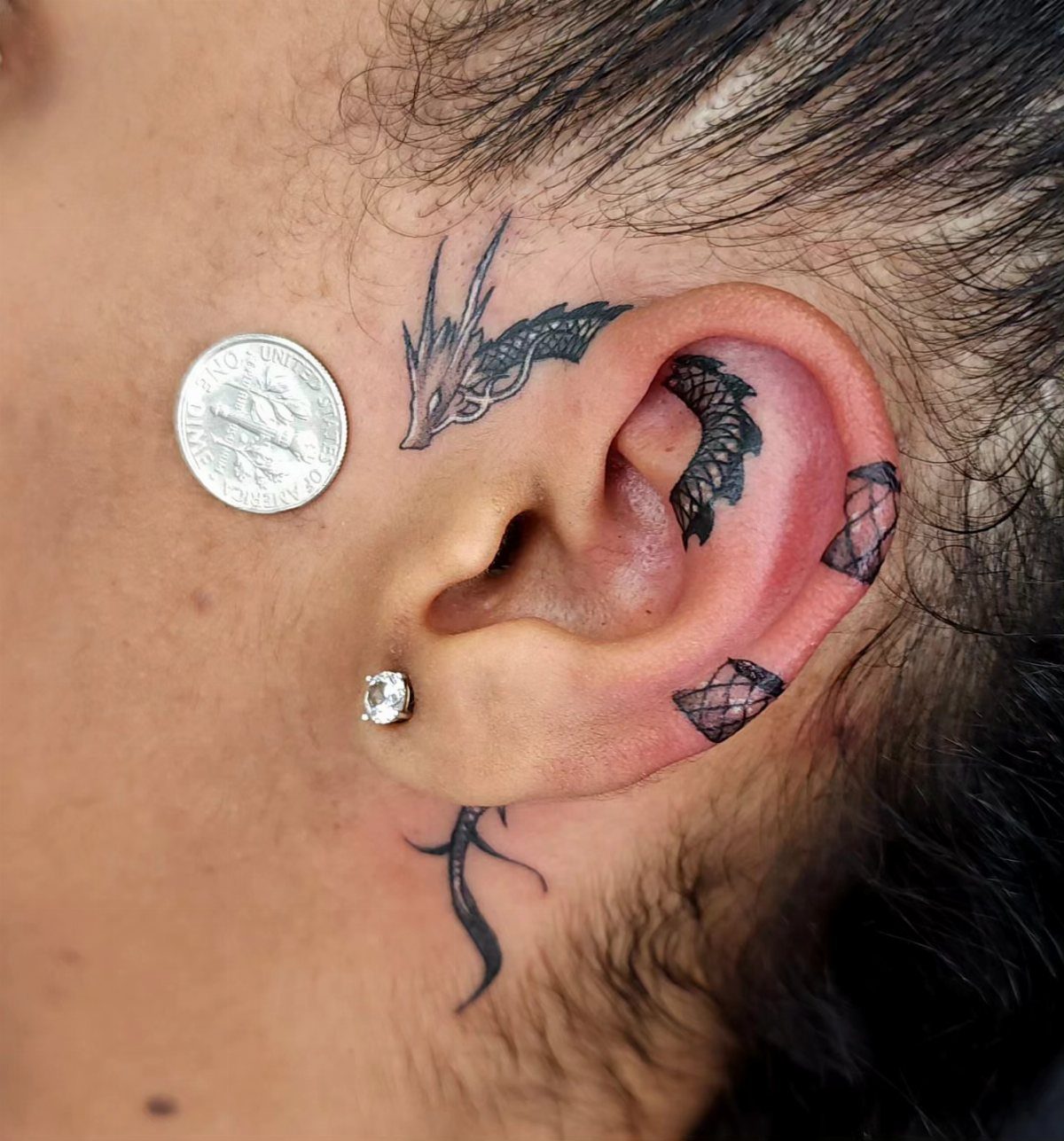 Small black dragon tattoo wrapping around a person's ear and cheek with a coin.