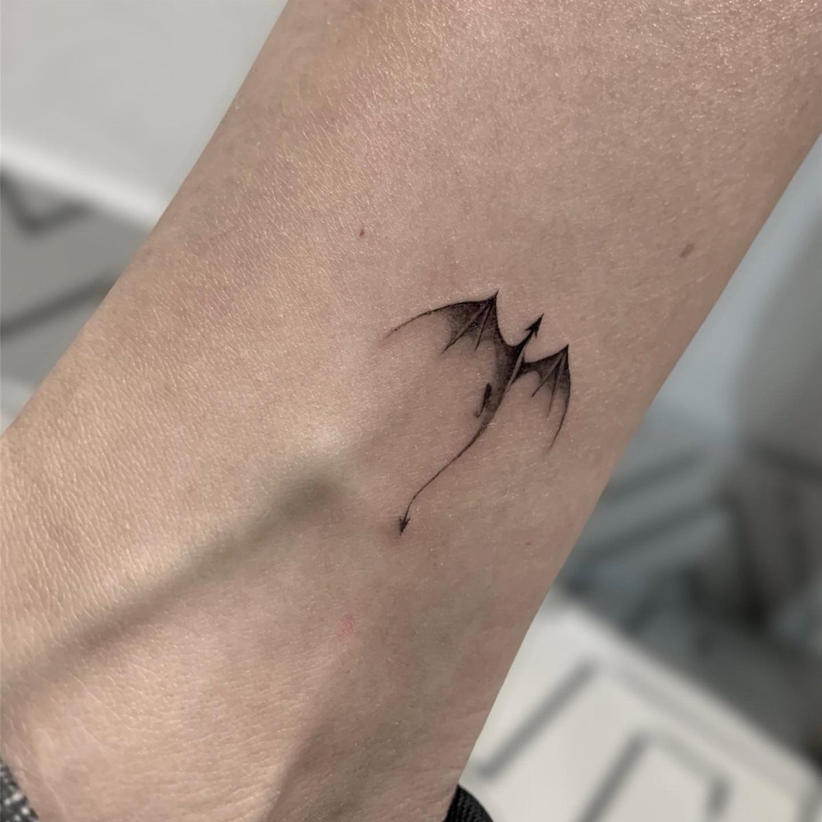 Minimalist black ink dragon tattoo with wings on a person's forearm.