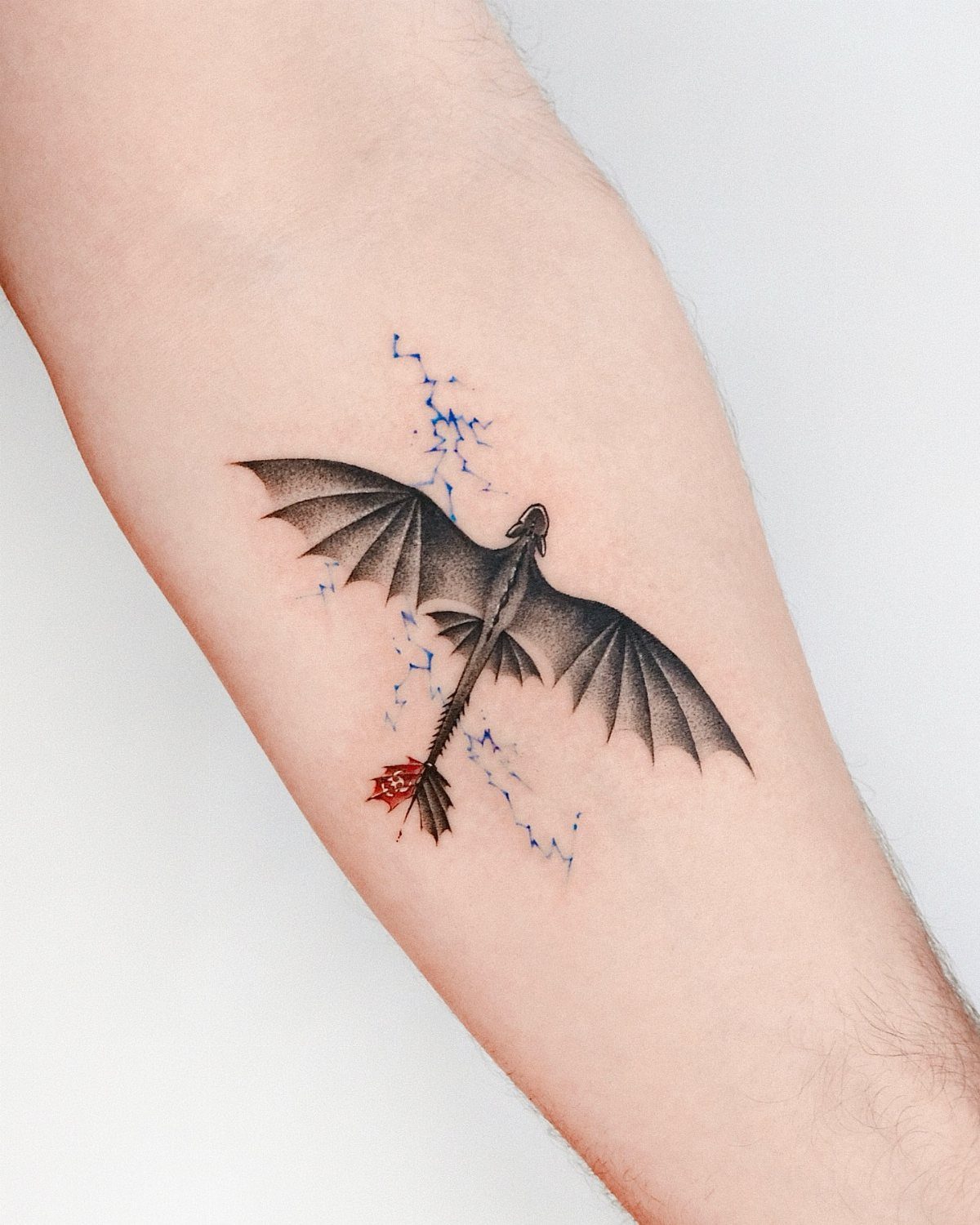 Toothless dragon tattoo with red tail fin and blue lightning on a forearm