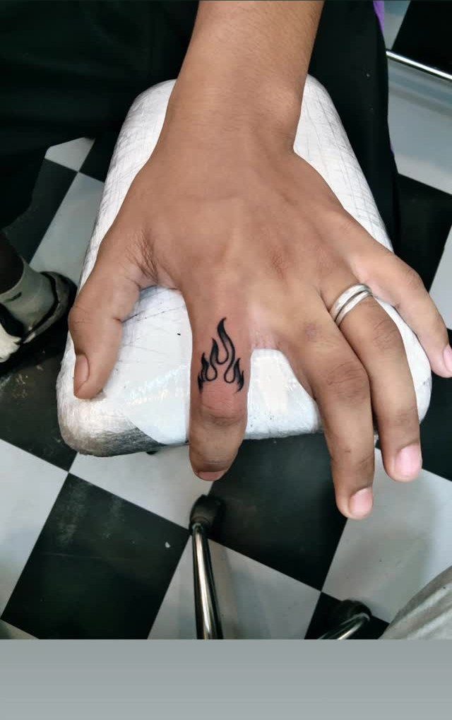 Small black flame tattoo on the middle finger of a person's hand.
