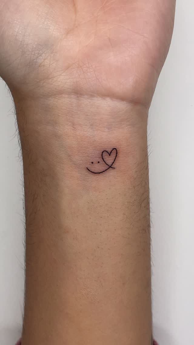 Small black heart tattoo with a curved line on a person's inner wrist