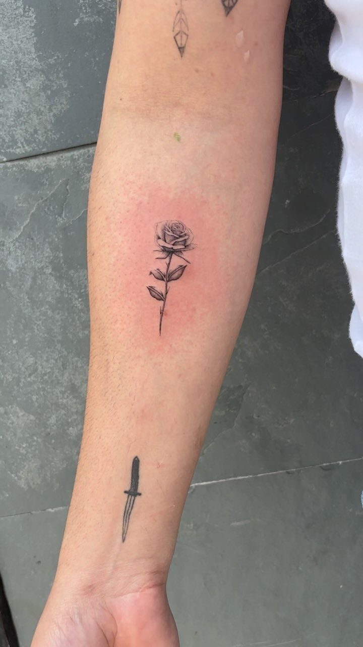 Small black ink rose tattoo and a dagger tattoo on a person's forearm.