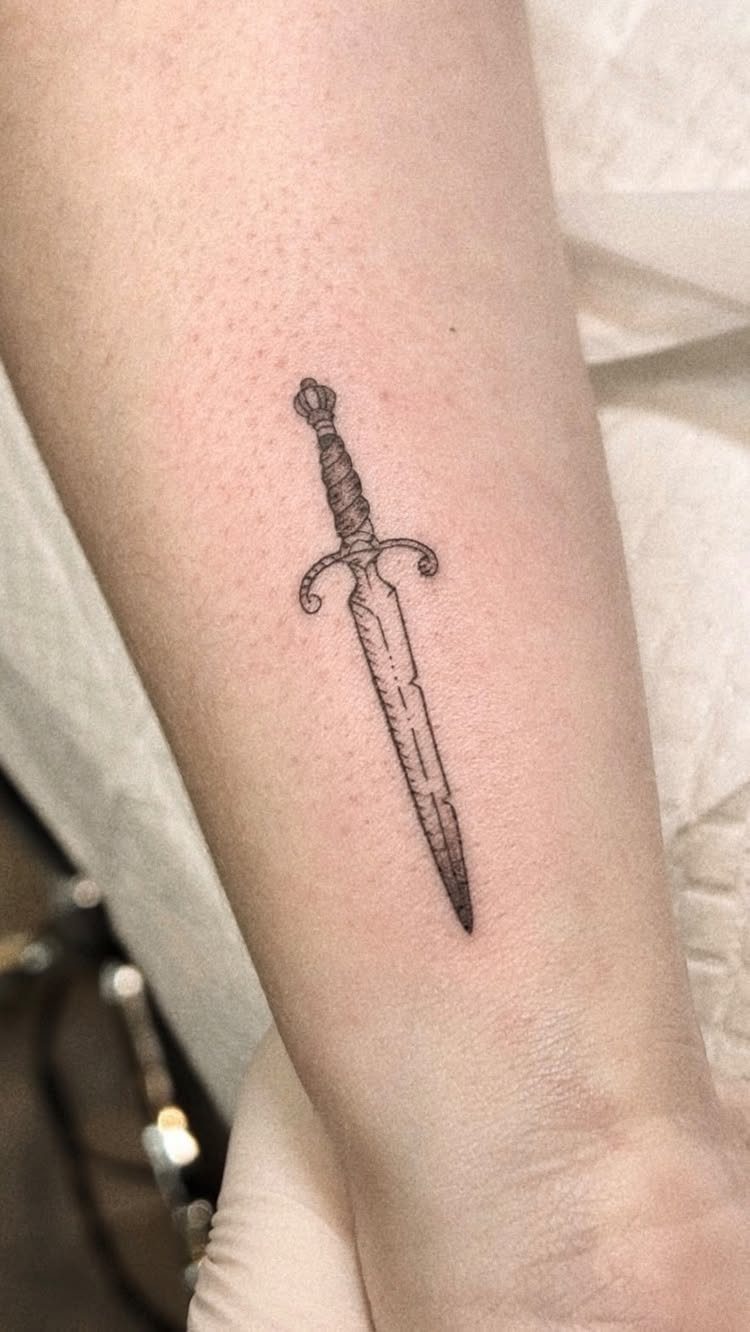 Small black ink dagger tattoo on a person's forearm.