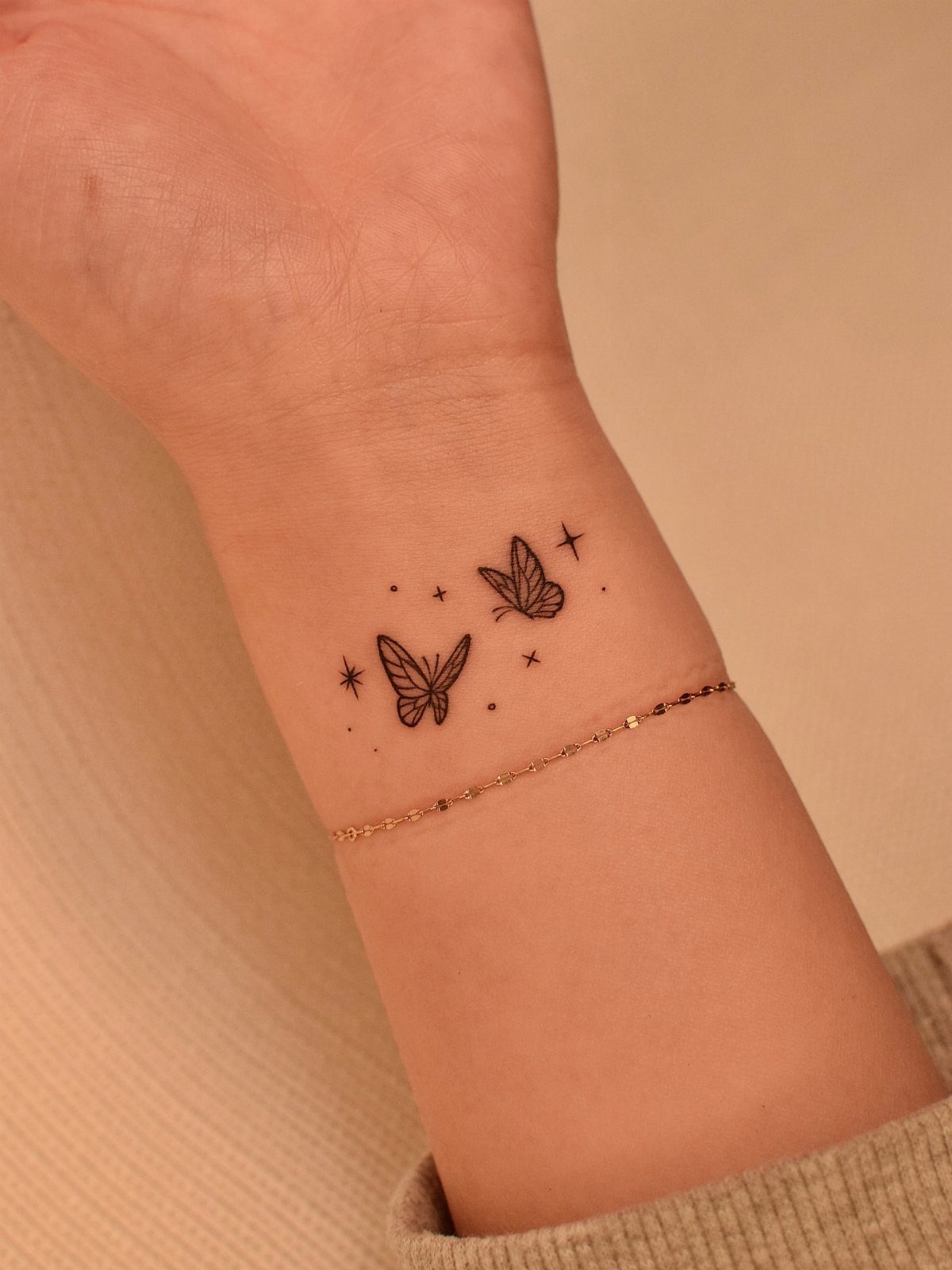 Two small black butterfly tattoos with stars on a person's wrist with gold bracelet
