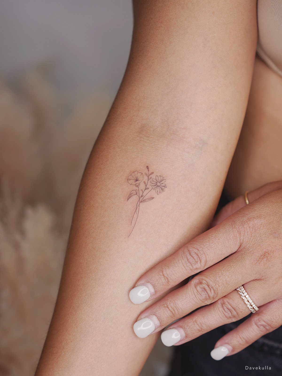 Small fine-line flower tattoo on a person's forearm with white manicured nails.