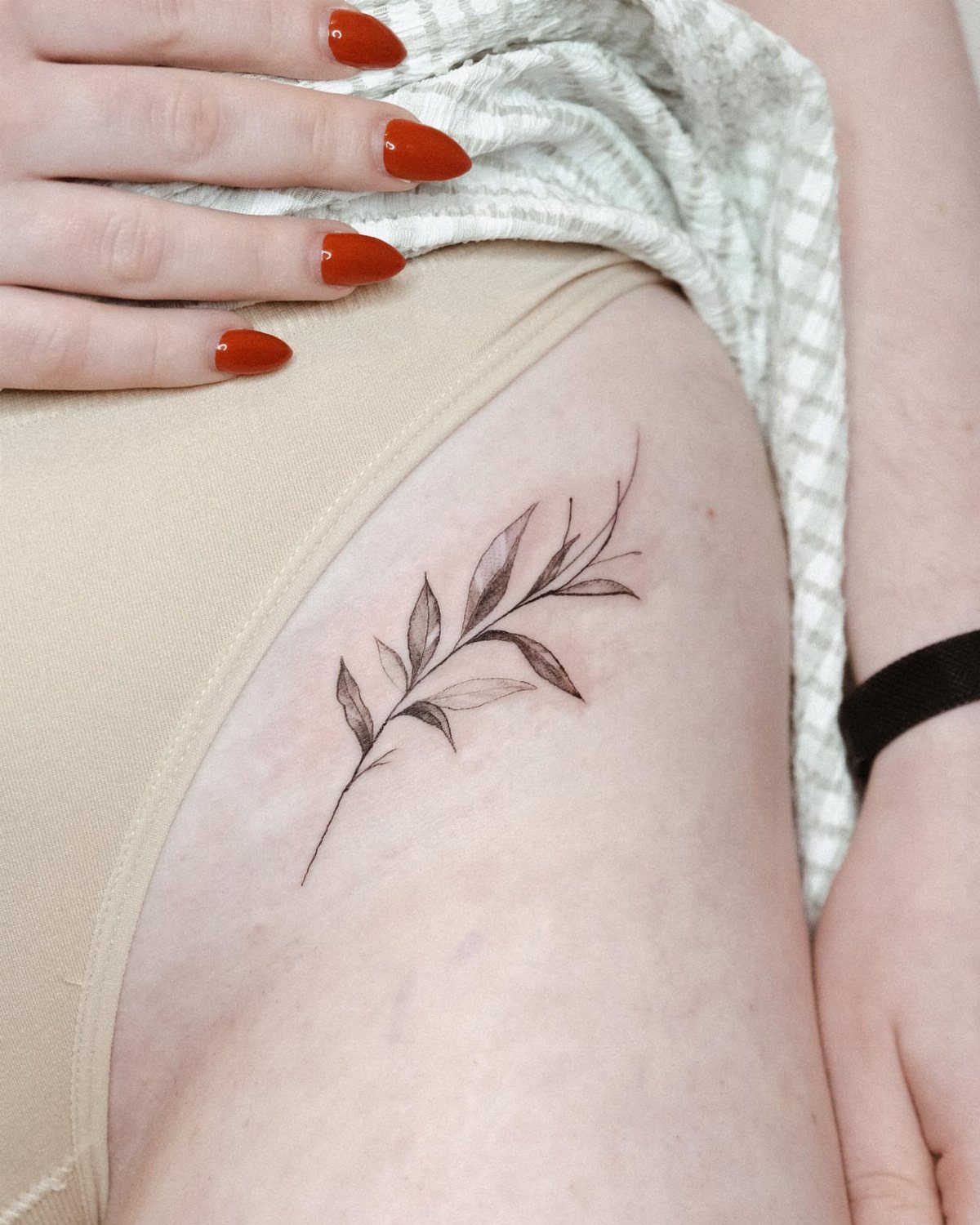 Small leafy twig tattoo on a person's hip with red nail polish.