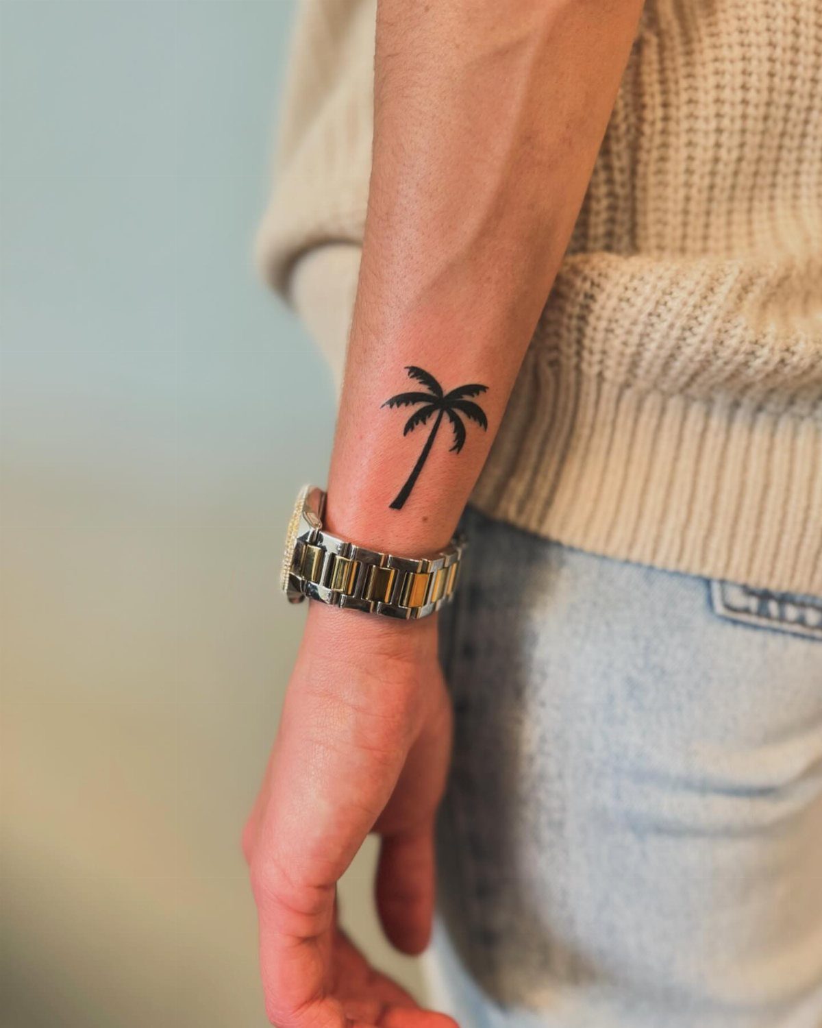 26 Small Pretty Tattoos for Butterflies, Stars, and More 2 Small black palm tree tattoo on a person's wrist next to a metal watch.
