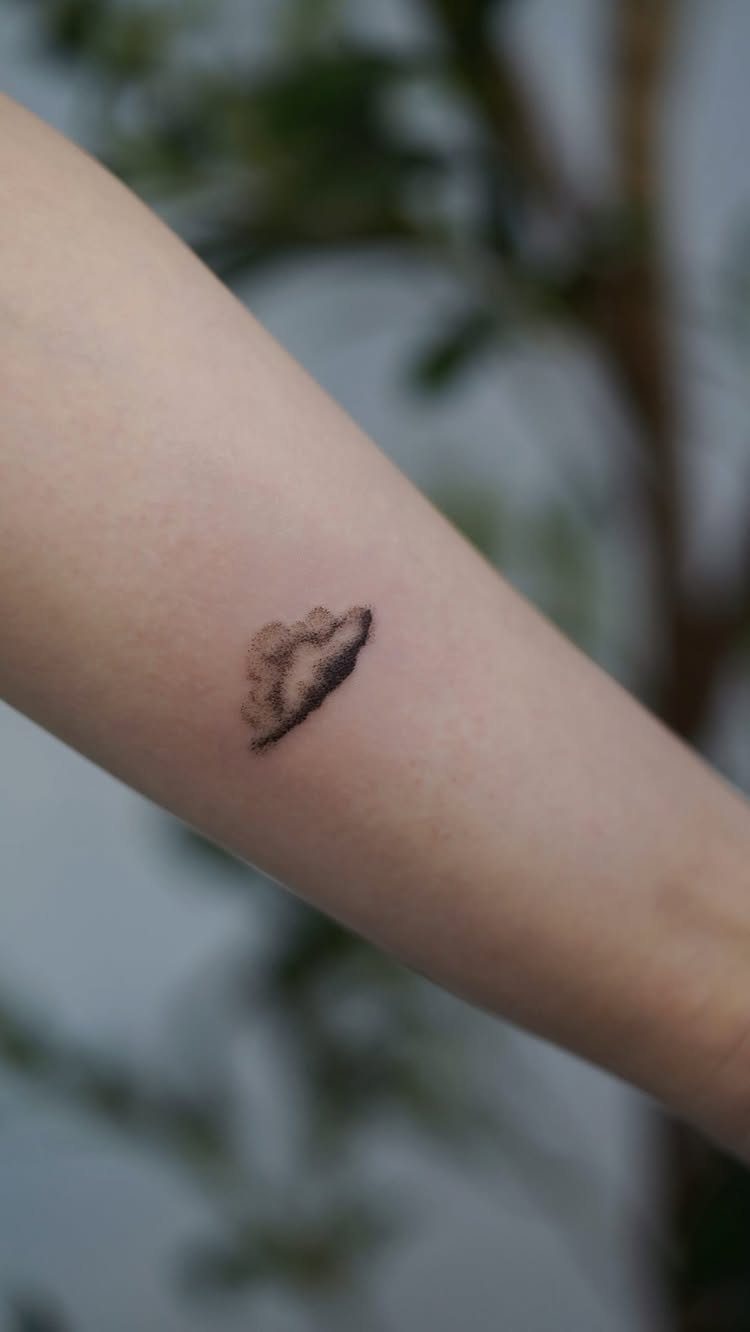 26 Small Pretty Tattoos for Butterflies, Stars, and More 26 Small black ink cloud tattoo on a person's forearm with blurred greenery background.