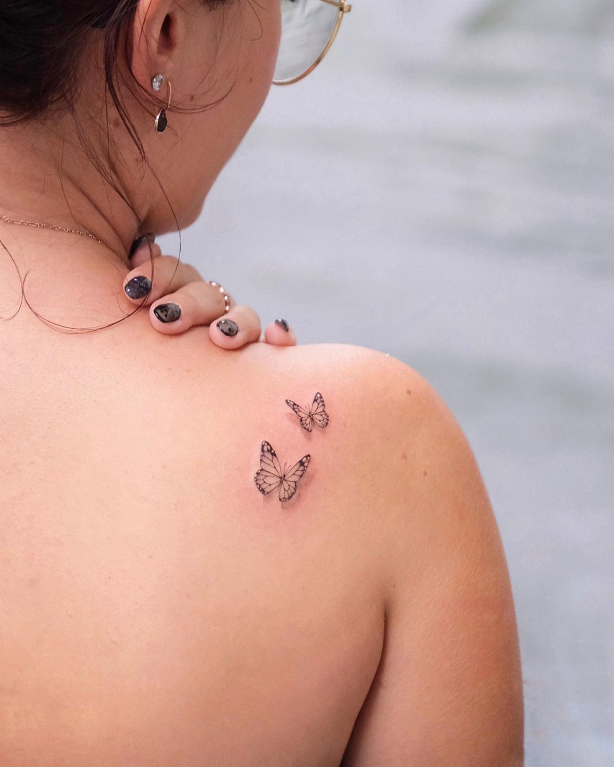 Two small fine-line butterfly tattoos on a person's upper shoulder and back.