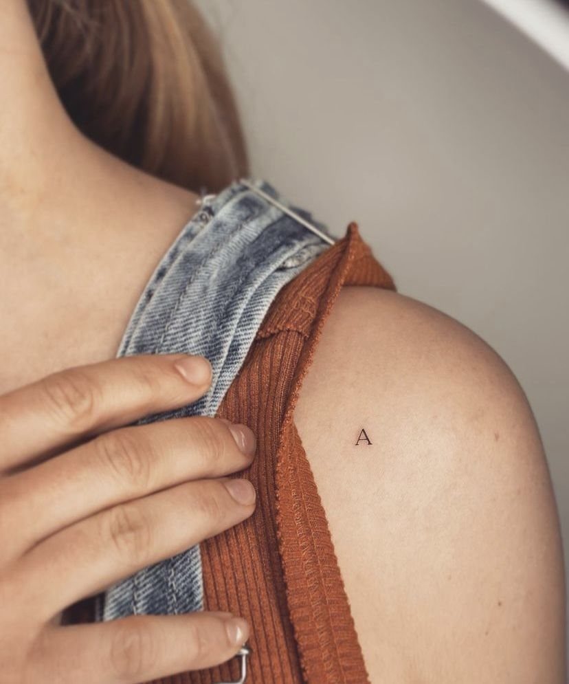Small black tattoo of the letter A on a person's shoulder