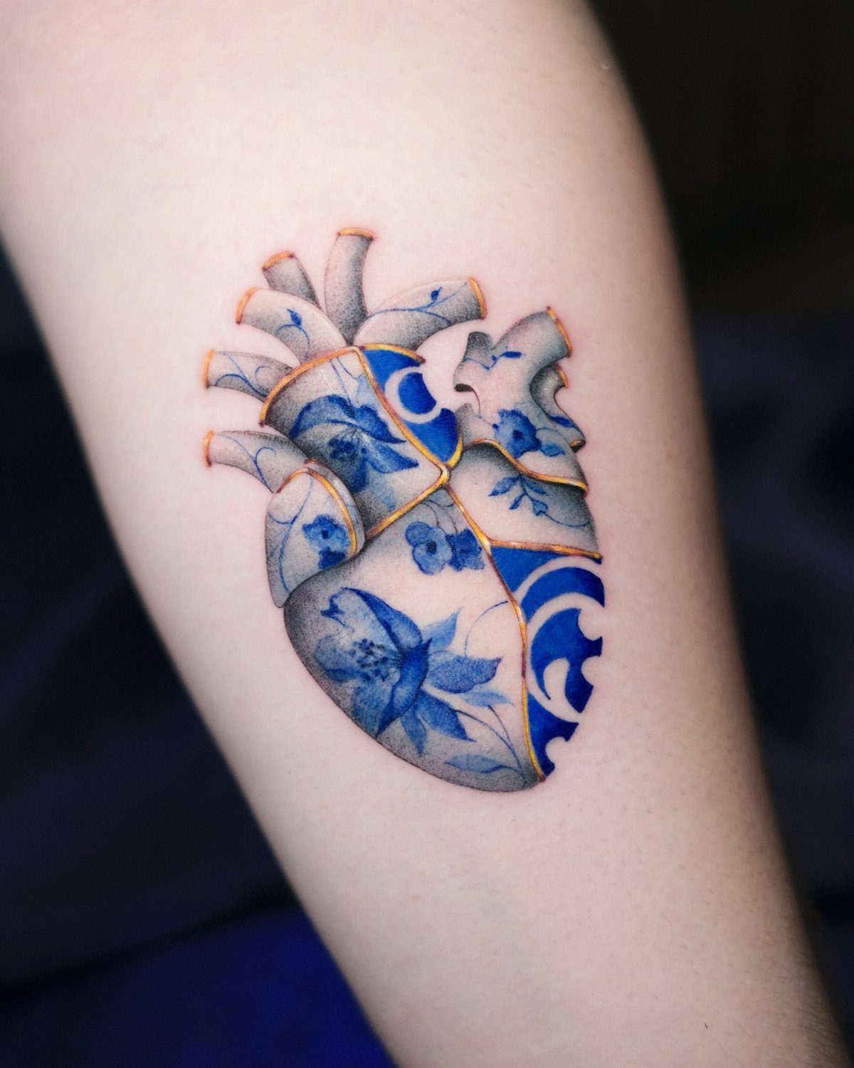 Anatomical heart tattoo on an arm featuring blue porcelain patterns and gold kintsugi cracks.