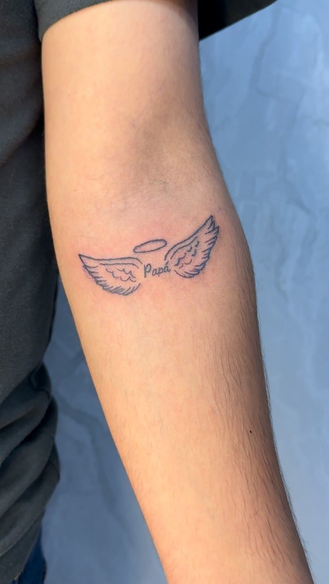 Angel wings tattoo with a halo and the word Papá on a forearm