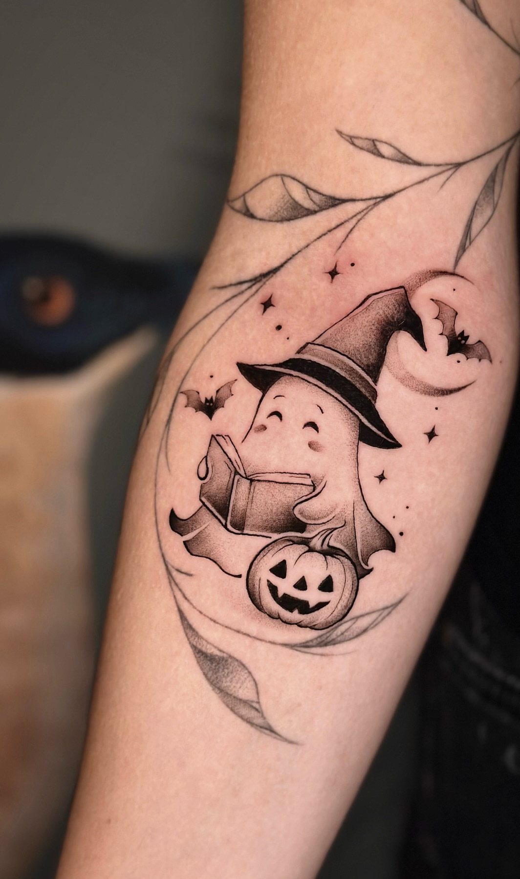 Cute ghost tattoo wearing a witch hat and reading a book on an arm