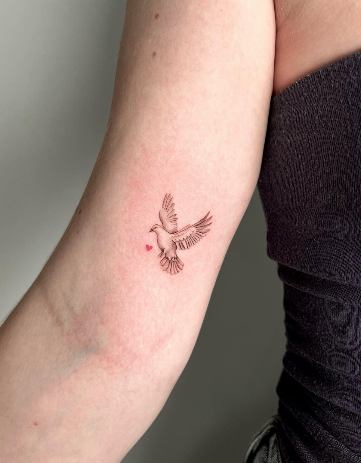 Small black ink dove tattoo with a tiny red heart on a forearm.