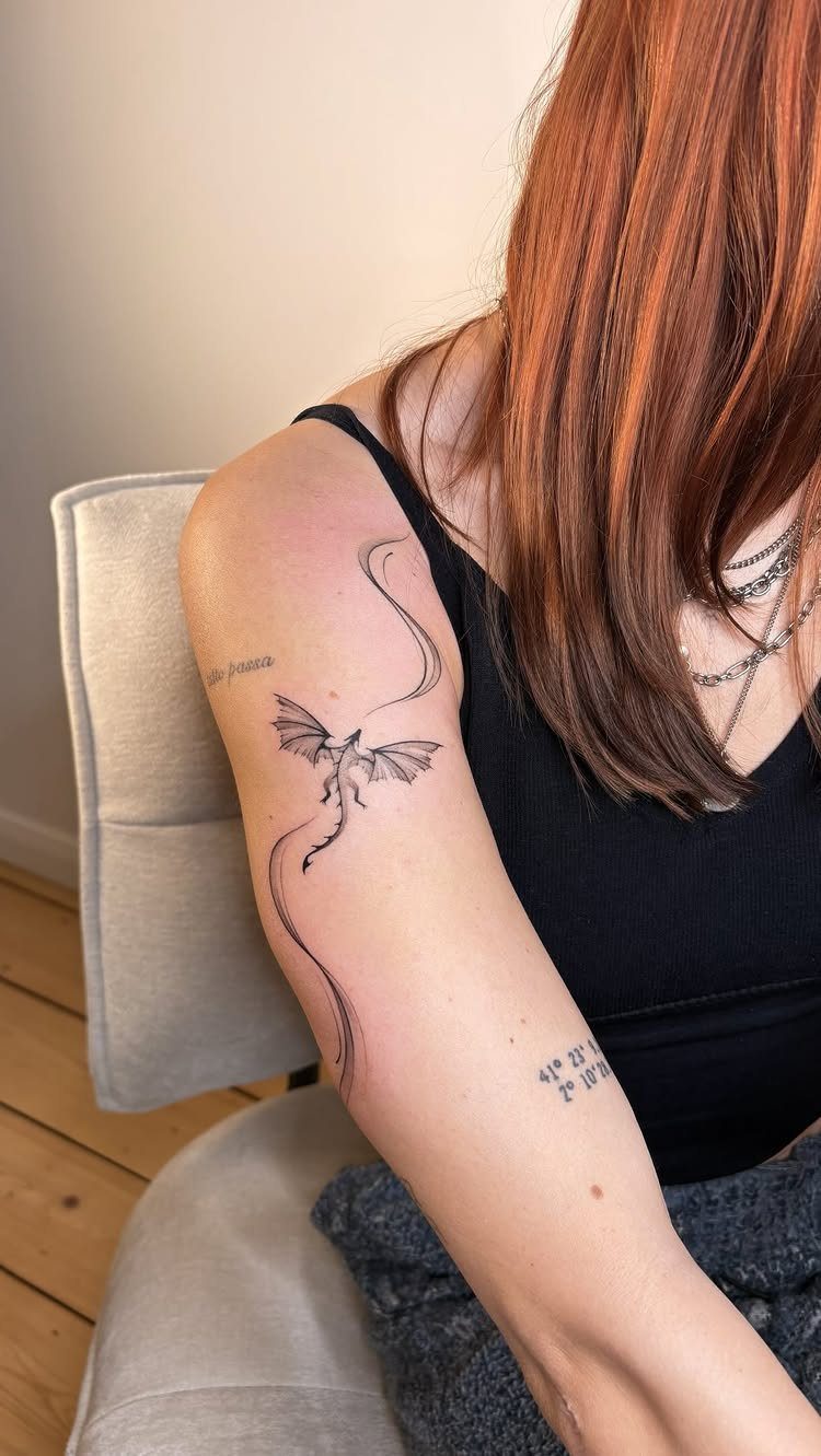 Small black ink dragon tattoo on a person's upper arm with red hair.