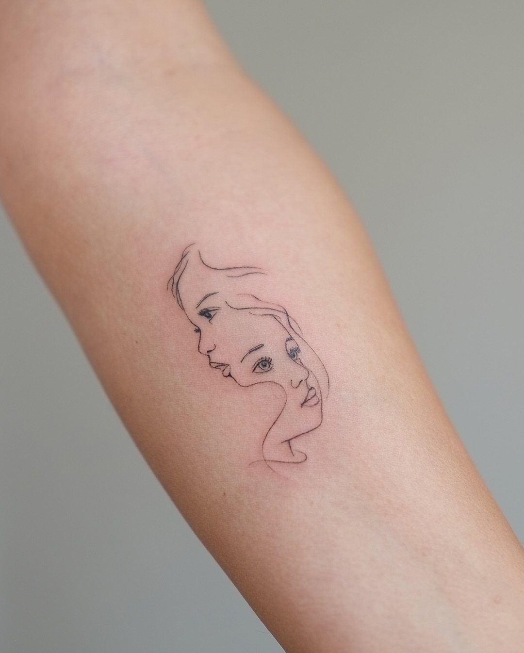 Minimalist line art tattoo of two overlapping female faces on a person's arm.