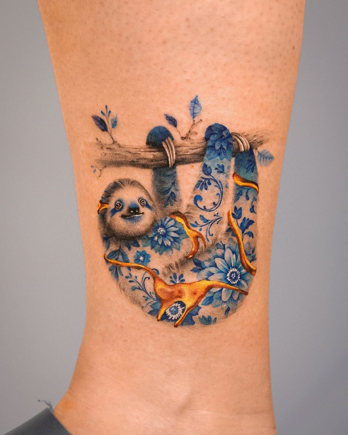 Sloth tattoo on arm with blue floral patterns and orange accents hanging from branch