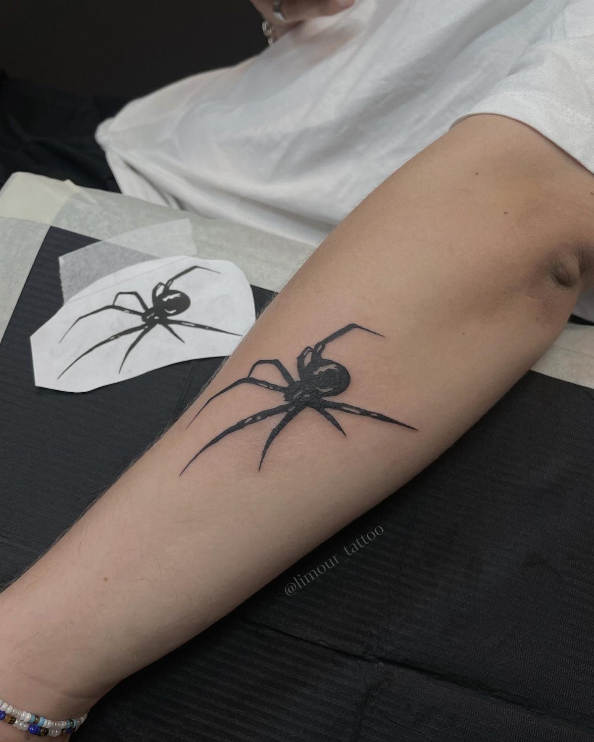 Black spider tattoo on a person's forearm next to a design sketch.