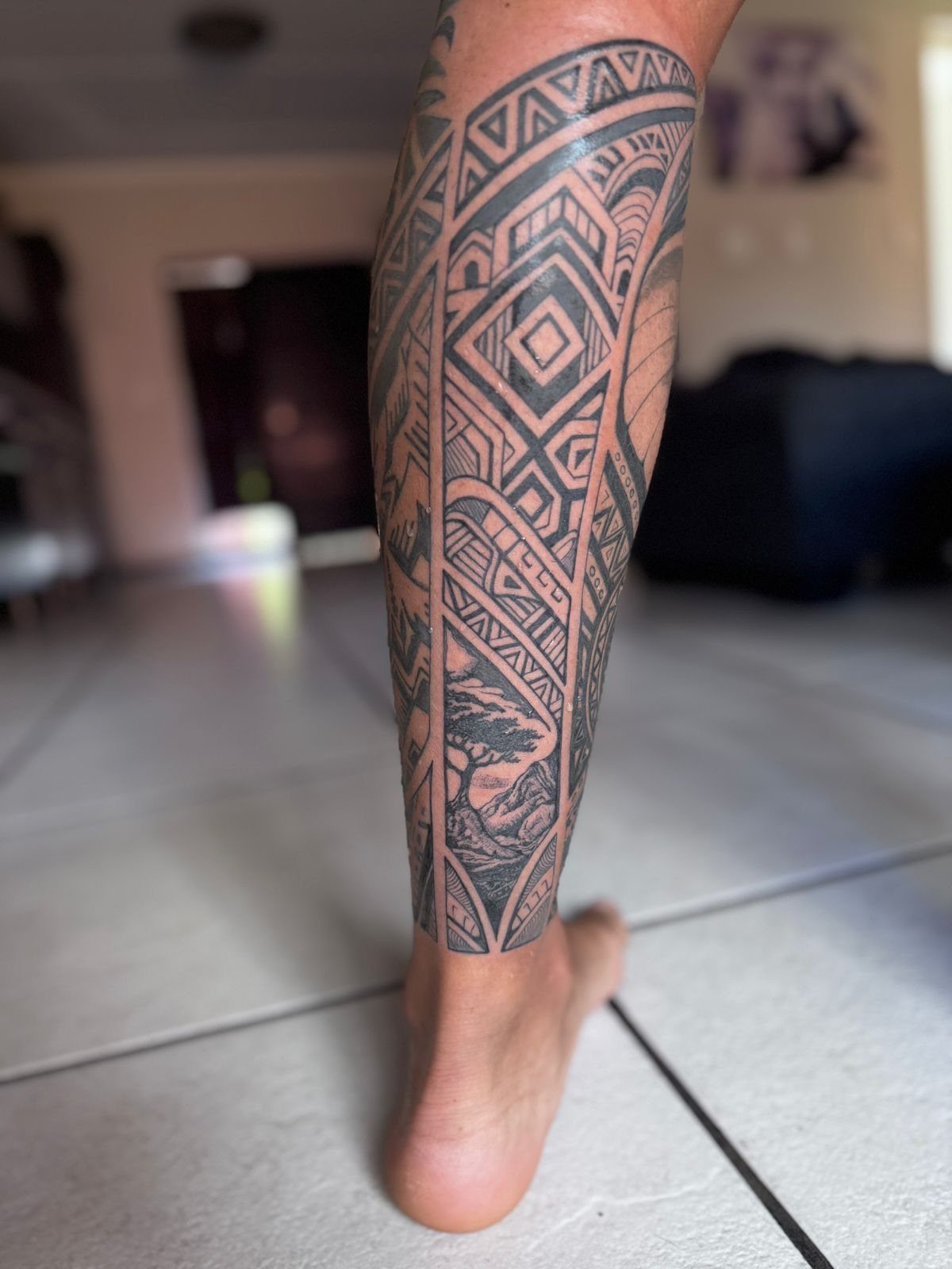 Lower leg with a detailed black geometric tribal tattoo and landscape scene.