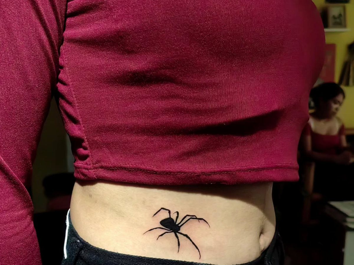 Black spider tattoo on a person's stomach with a maroon cropped shirt.
