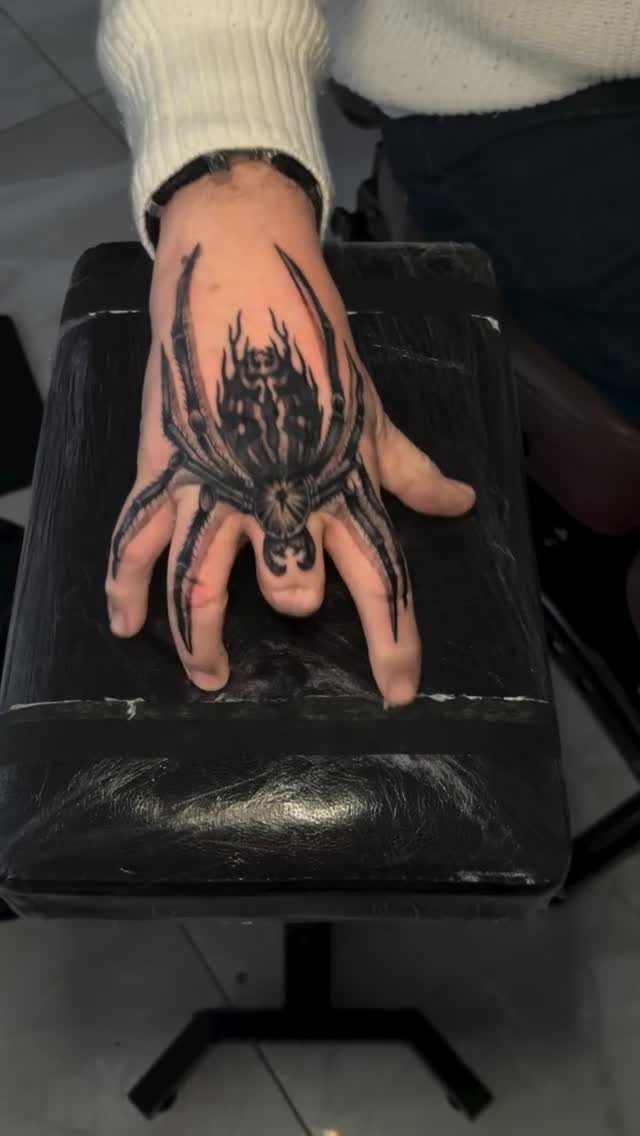 Black ink spider tattoo on a hand with legs extending over the fingers