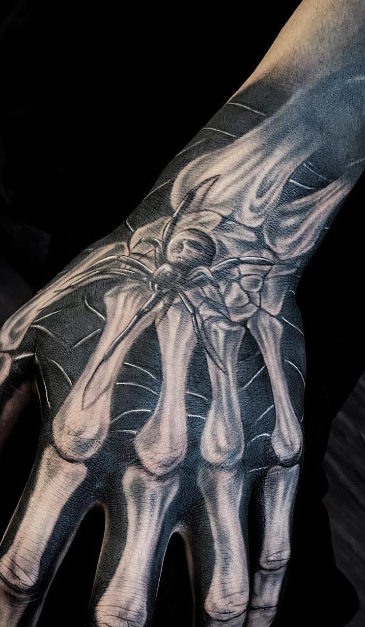 Spider tattoo on a web over skeletal hand bones in black and grey ink.