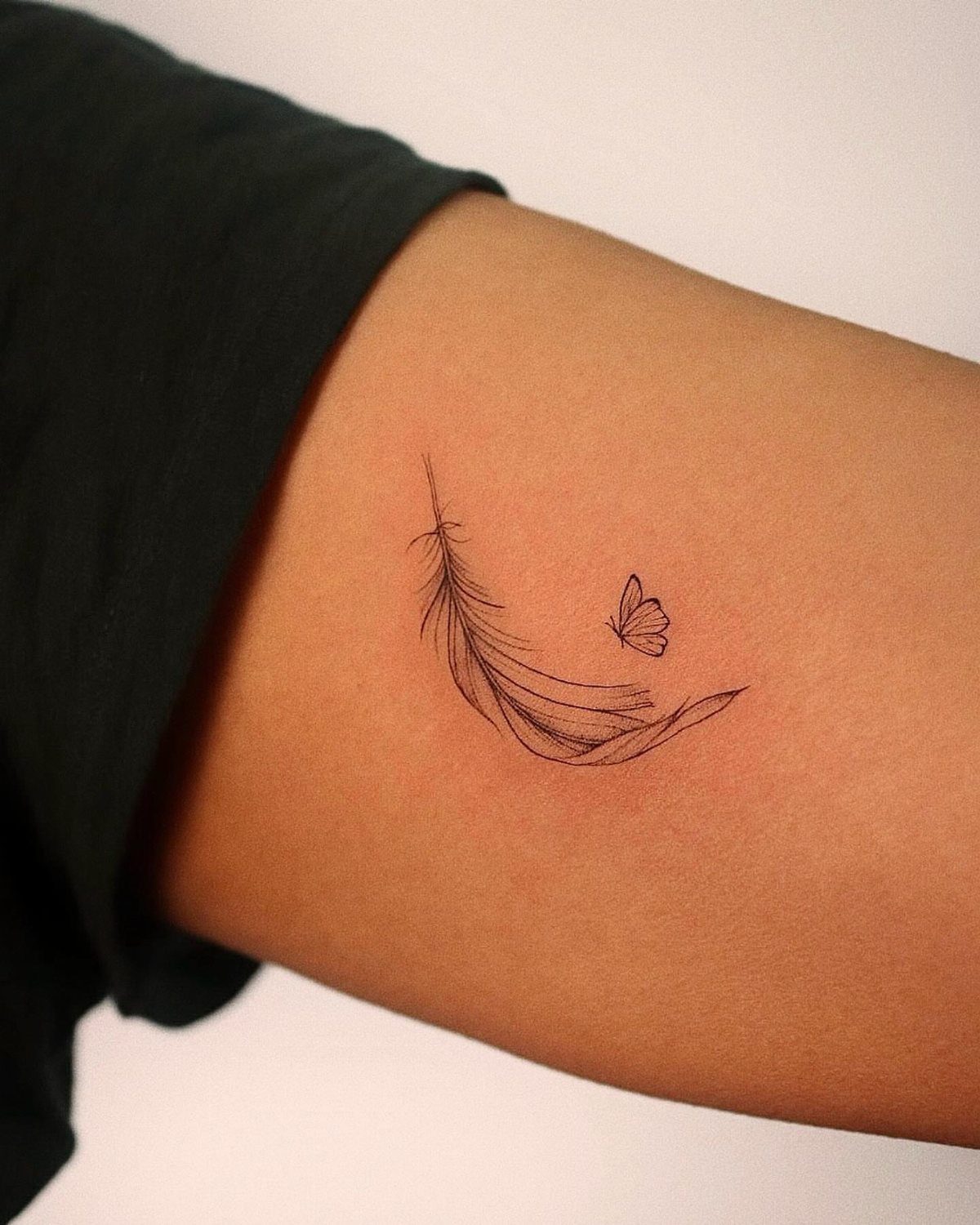 Fine line tattoo of a curved feather and small butterfly on a person's arm