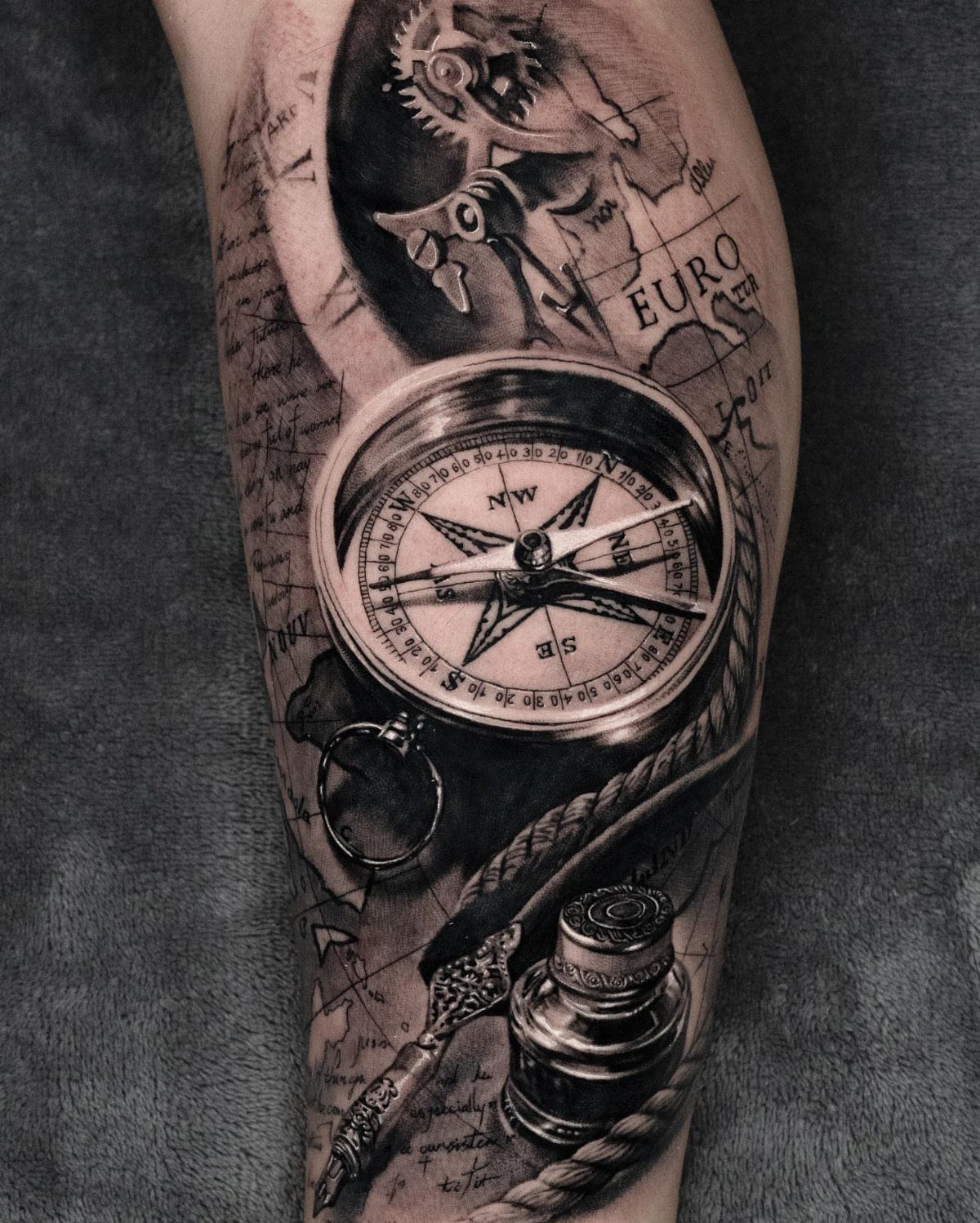 Detailed black and grey tattoo of a compass, ink bottle, and map on arm
