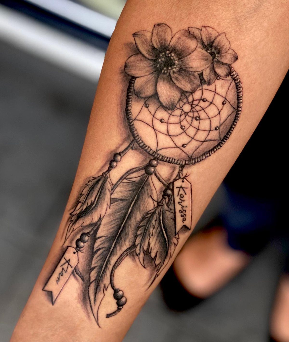 Dream catcher tattoo on an arm with flowers, feathers, and name banners.