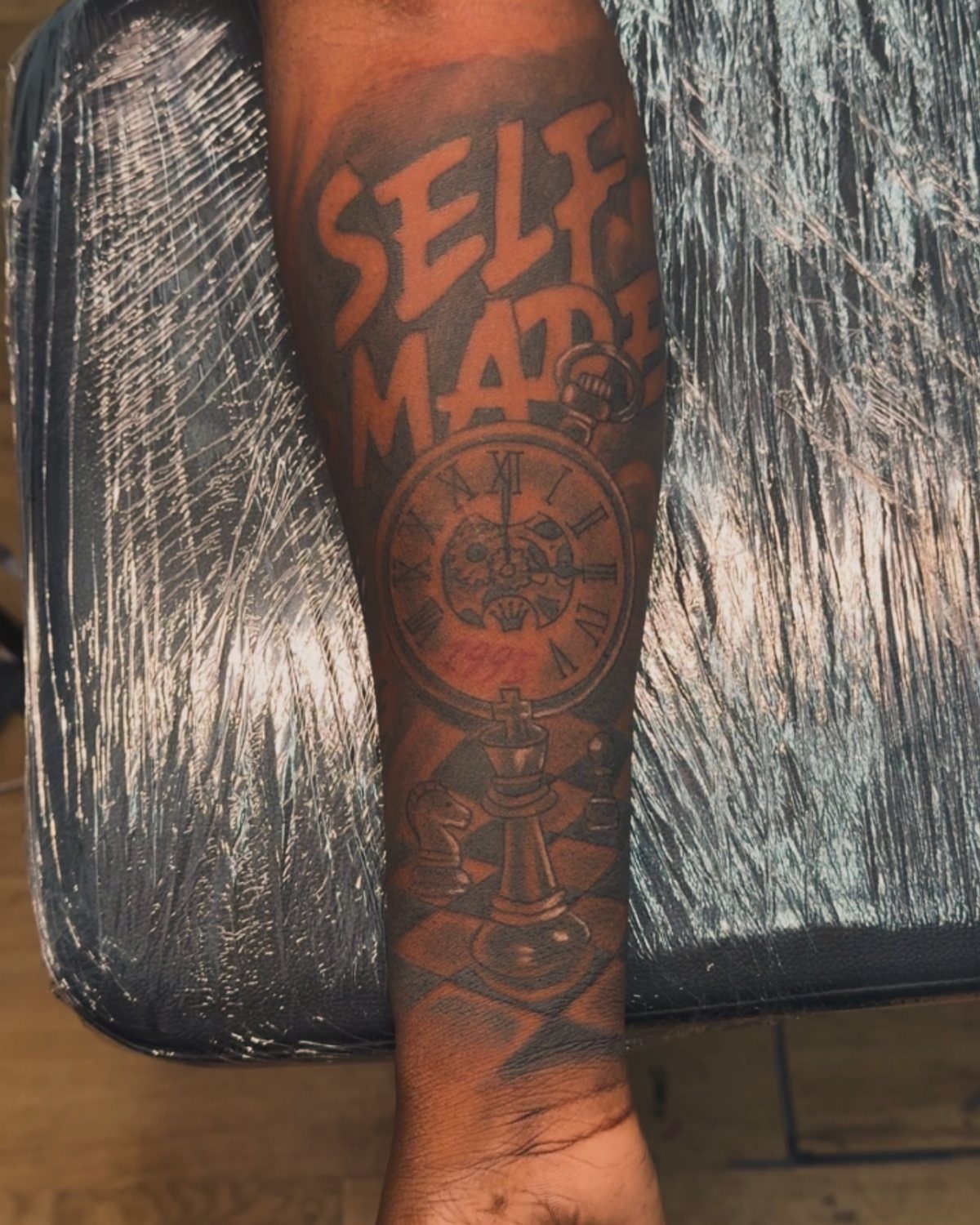 Forearm tattoo featuring the words SELF MADE, a clock, and a chess piece.