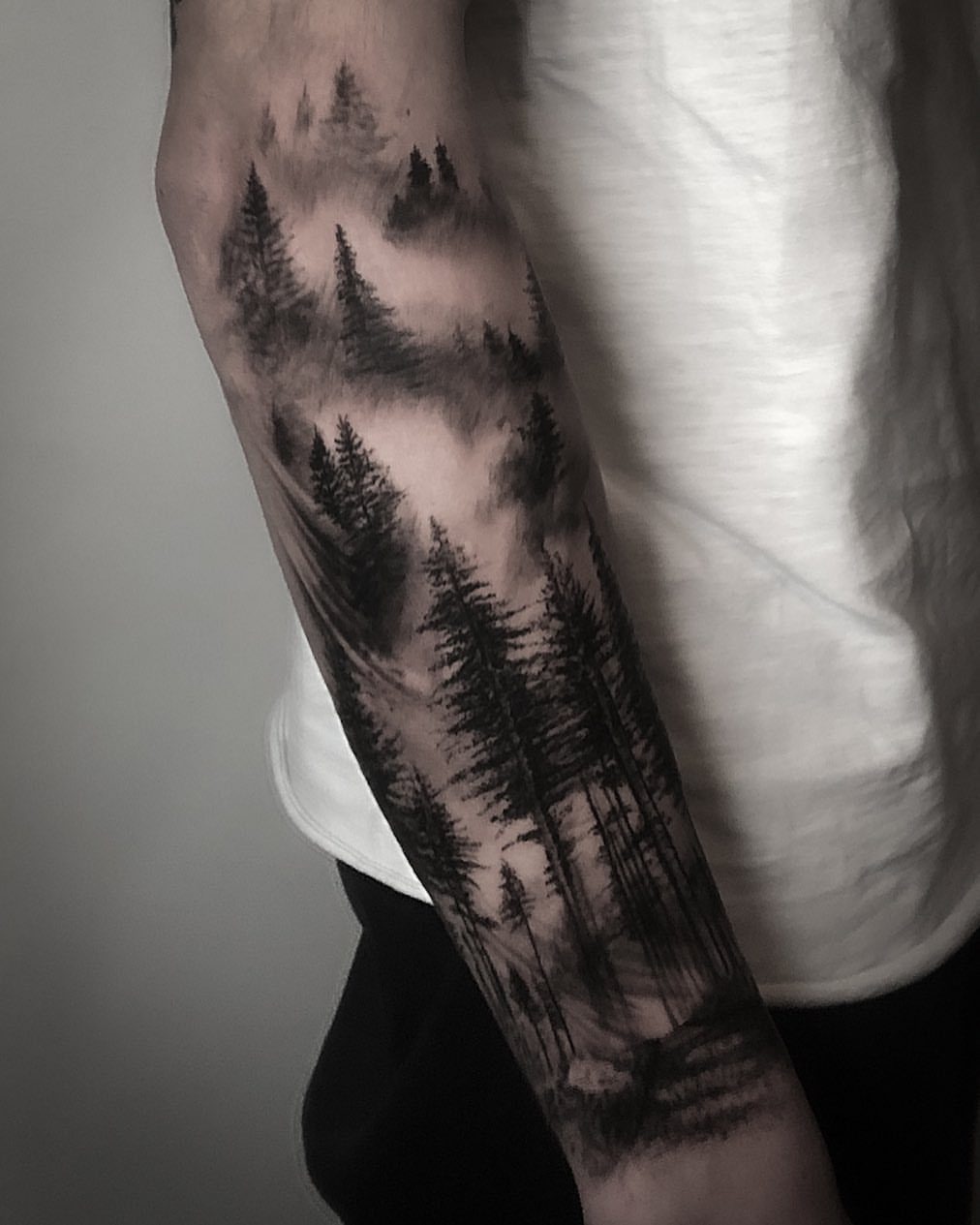 Forearm tattoo of a misty pine forest landscape in black and grey ink.