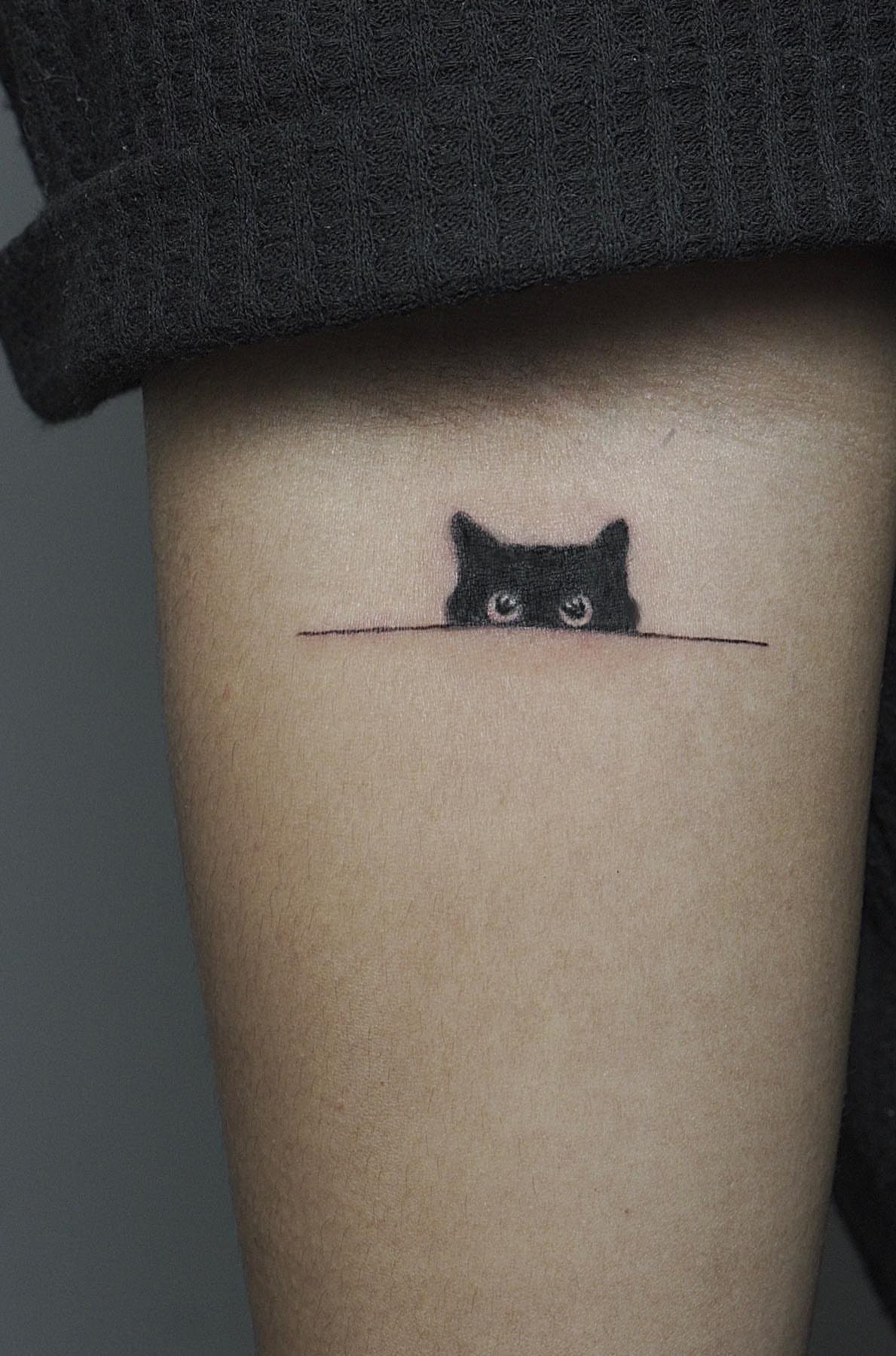 Small black cat tattoo peeking over a line on a person's arm.