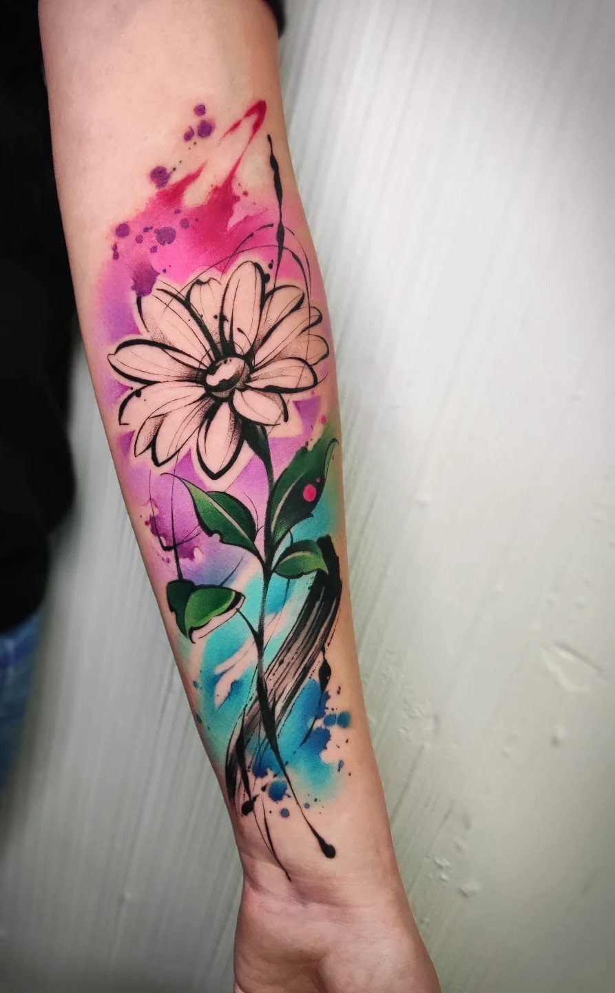 Watercolor floral tattoo of a white daisy with pink and blue splashes on forearm