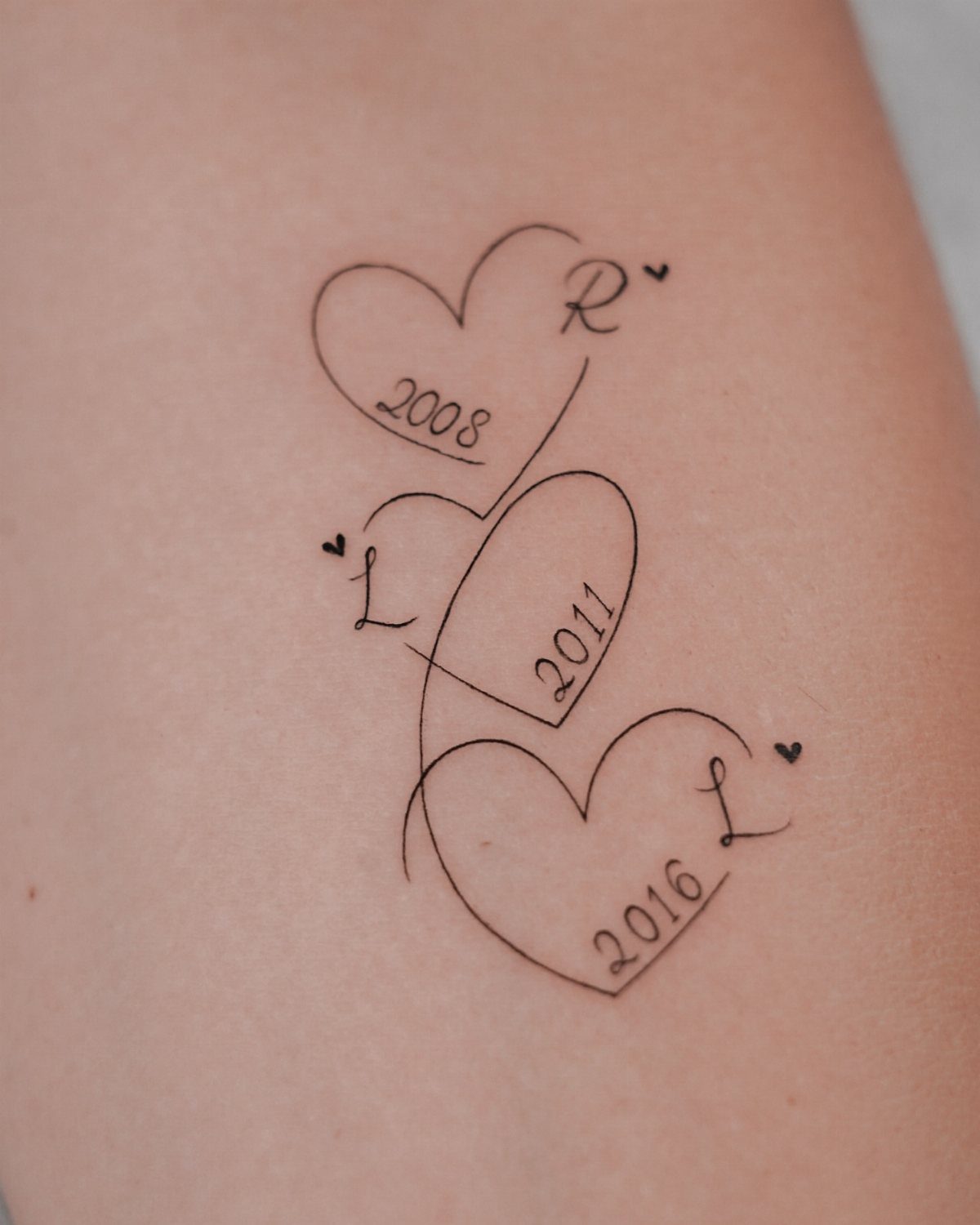 Three heart tattoos with initials and birth years on a person's skin