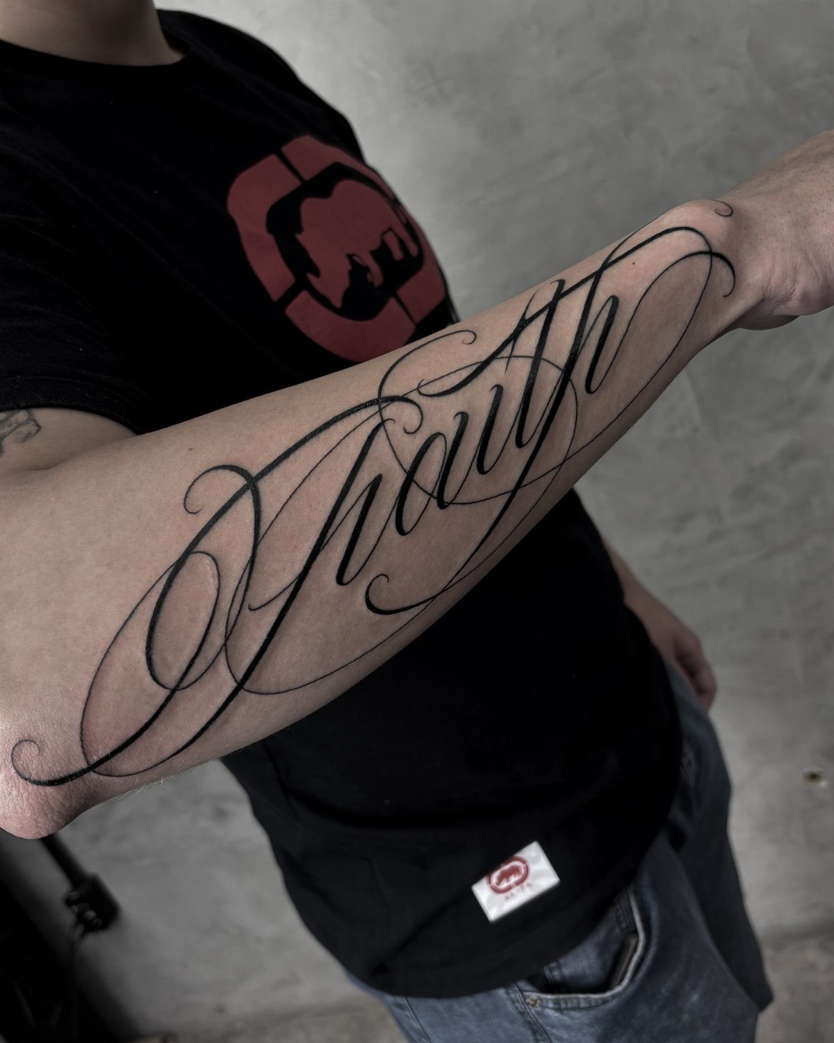 Forearm tattoo of the word Faith in elegant, flowing black ink calligraphy.