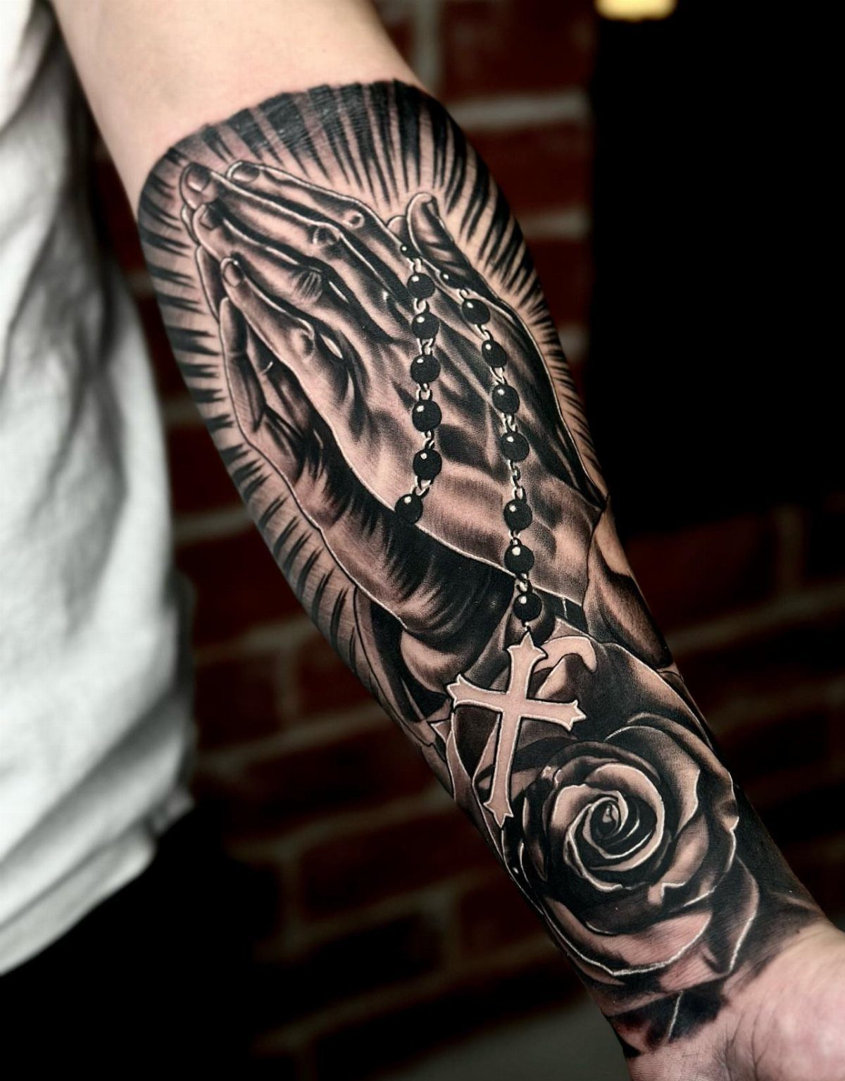 Praying hands tattoo with a rosary and rose on a person's forearm.