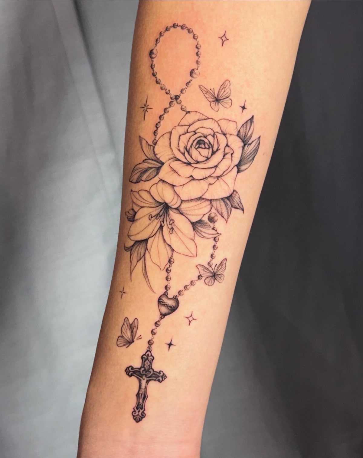 Rosary bead tattoo with a rose, lilies, and a crucifix on a forearm.