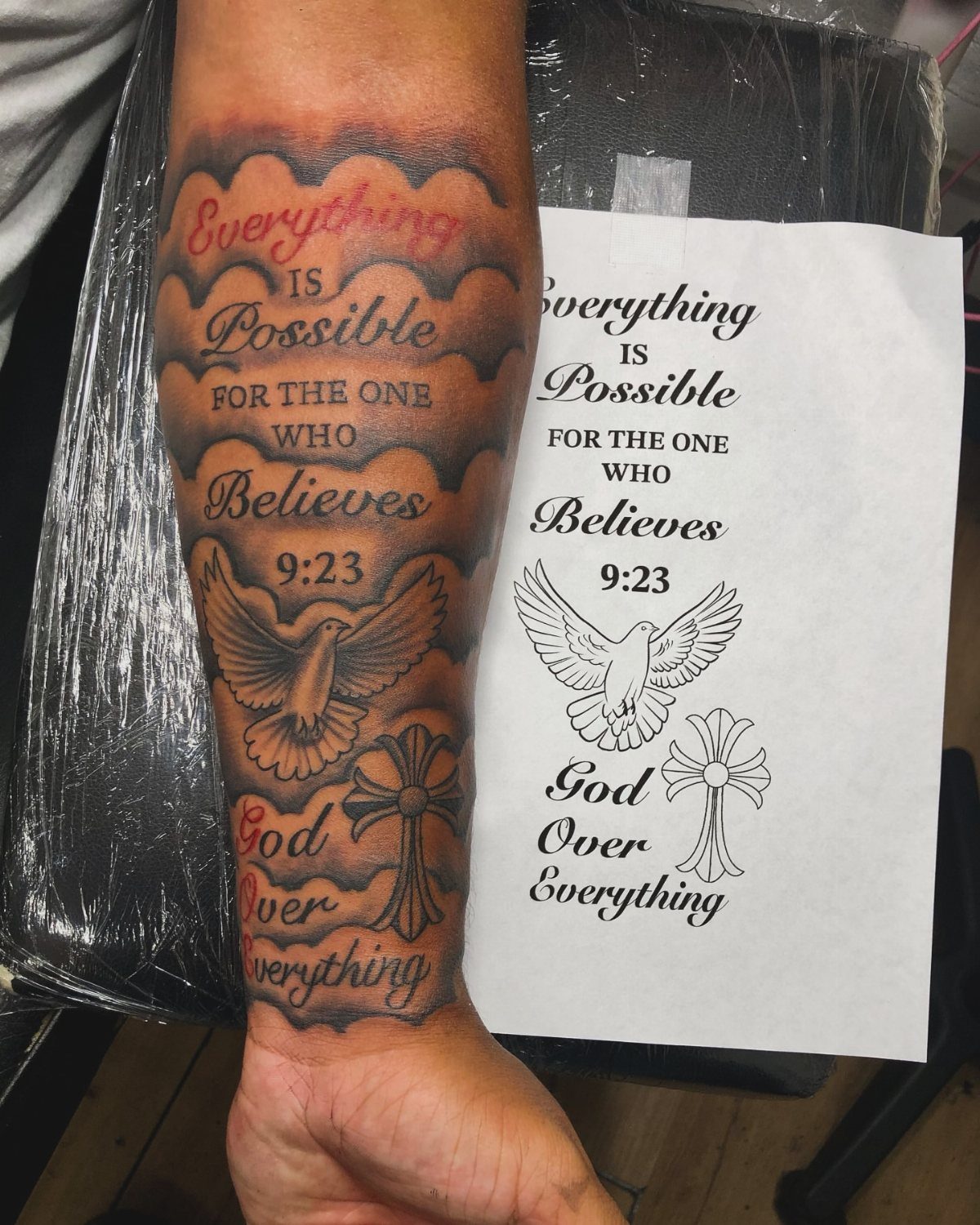 Forearm tattoo with biblical scripture, a dove, and a cross next to design paper.
