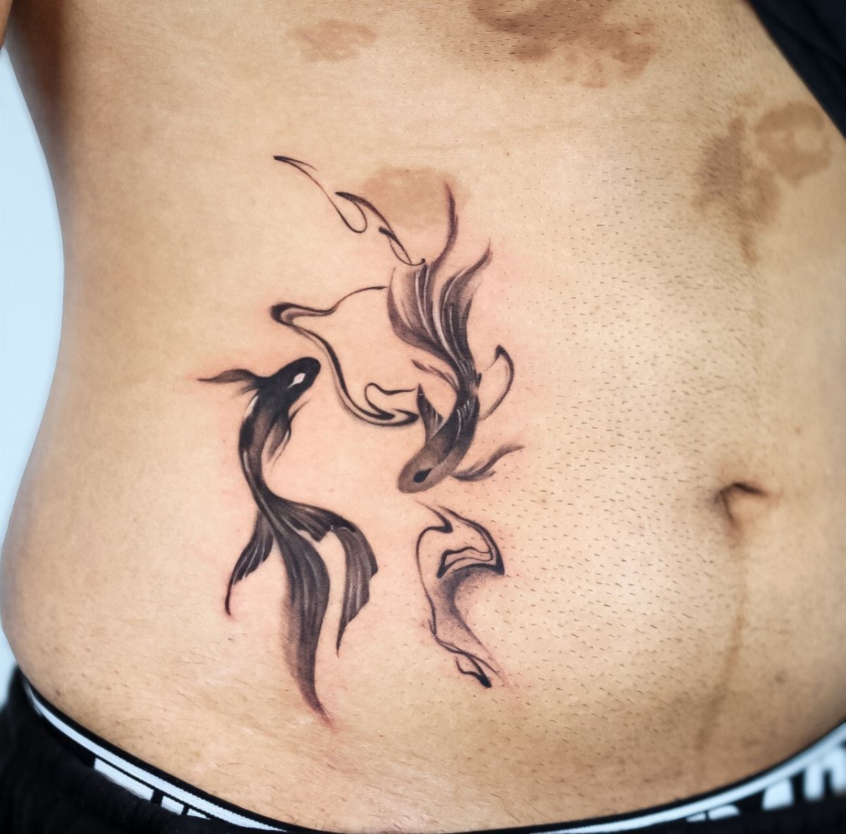 Tattoos Around Belly Button: 8 Best Ideas to Consider 2 Two black koi fish tattoos swimming in a circle on a person's abdomen.