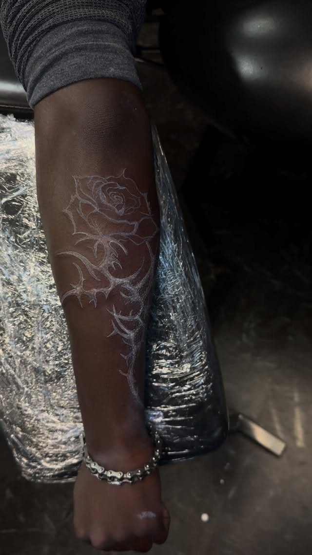 White ink rose tattoo on a person's dark-skinned forearm resting on plastic wrap.
