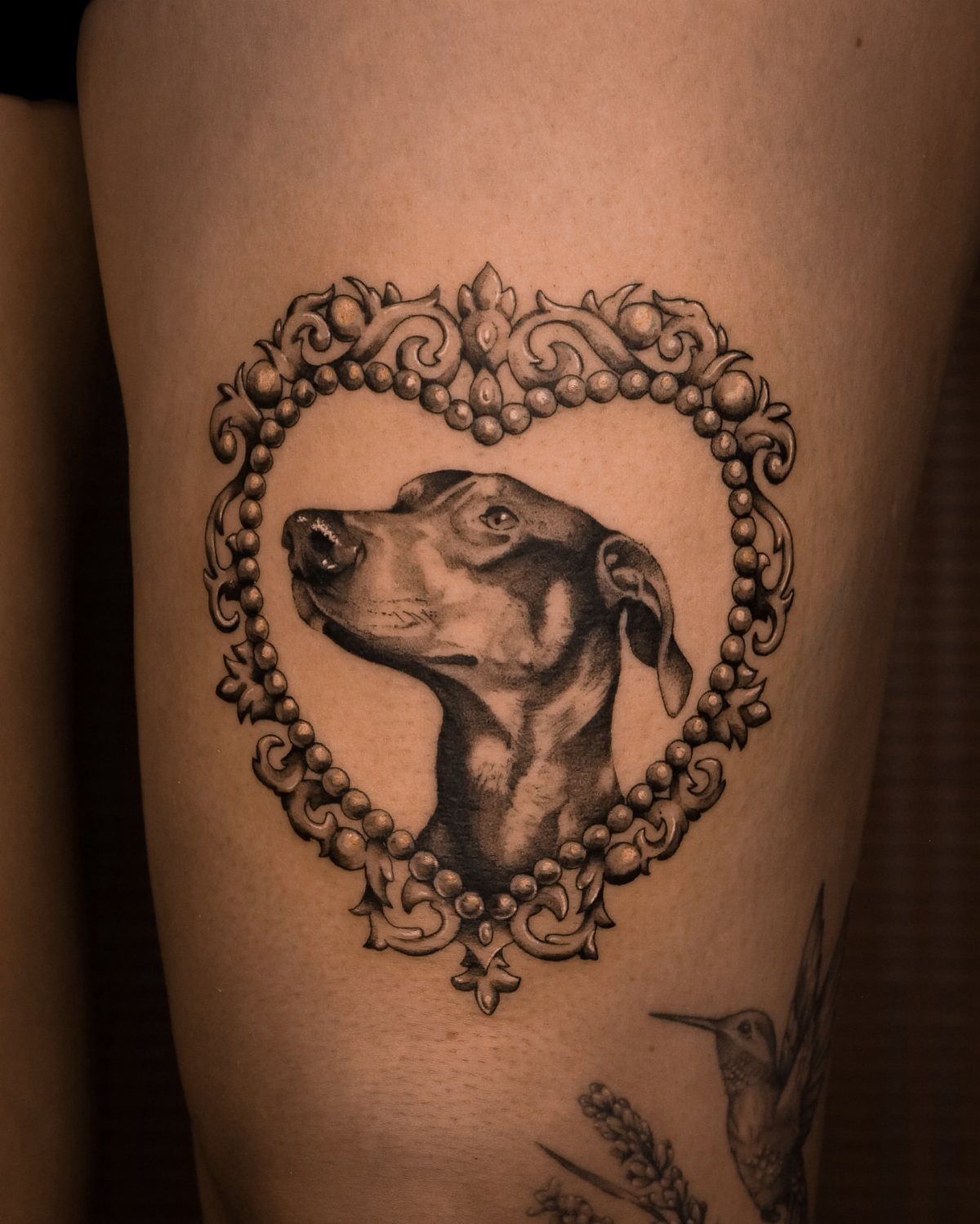 Dog portrait tattoo inside an ornate heart-shaped frame on a person's thigh.