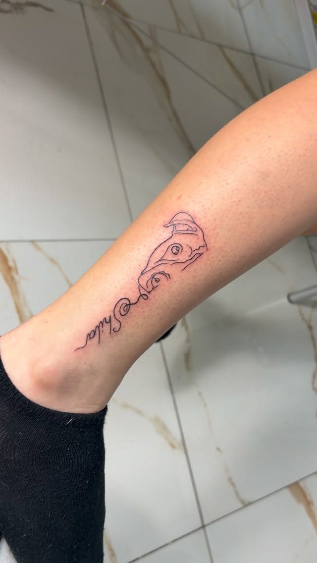 Line-art dog head tattoo with the name Shila on a person's lower leg