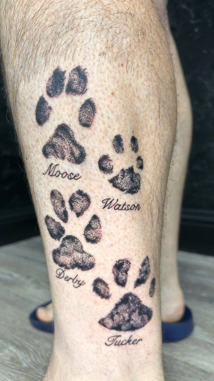 Four black paw print tattoos with names on a person's lower leg.