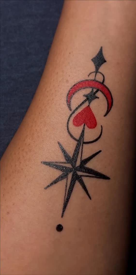 Tattoo on an arm featuring a red crescent moon, heart, and black compass star.